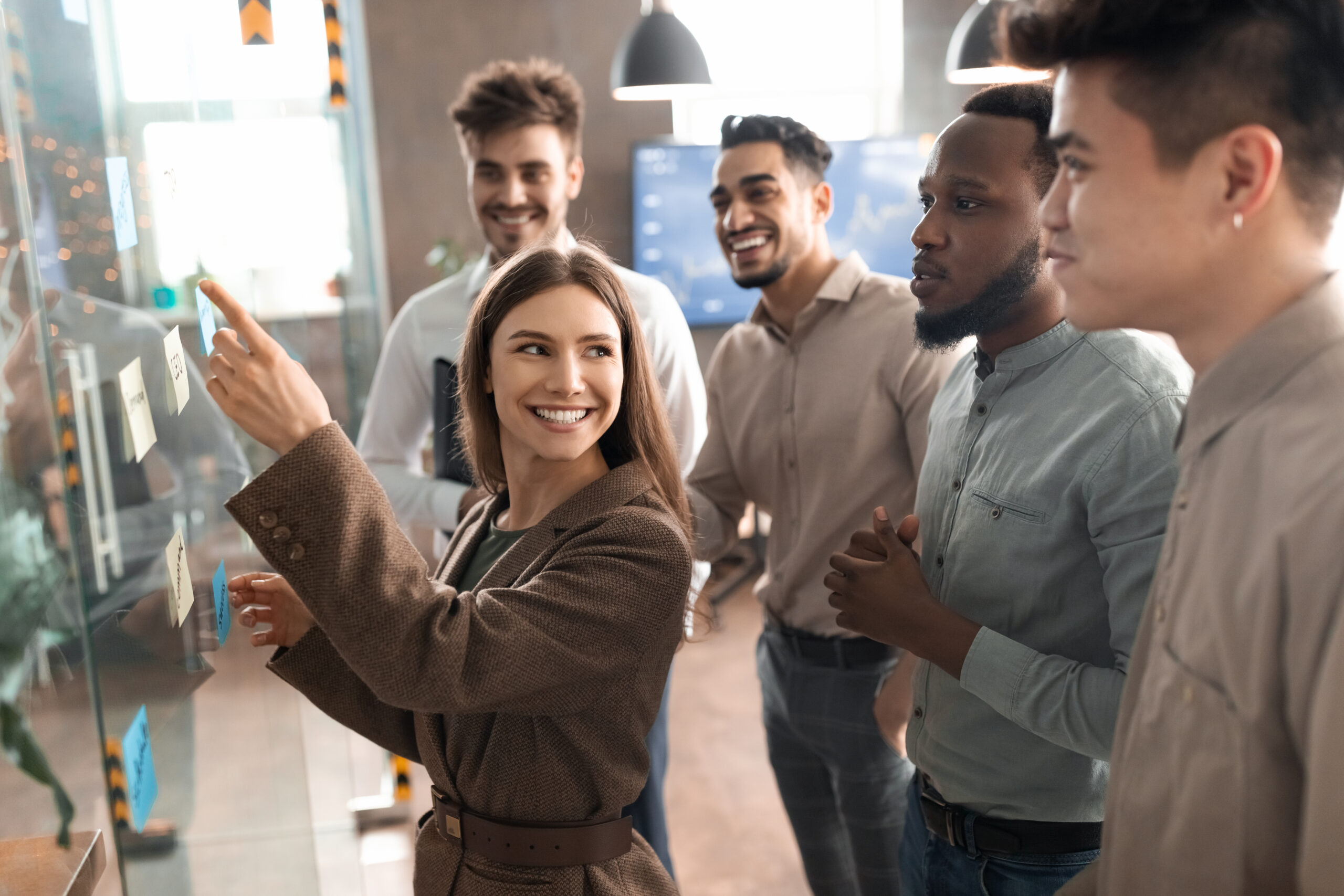 Teamwork Concept. Creative group of smiling professional businesspeople brainstorming, wonan pointing at sticky post-it paper notes on glass wall, discussing ideas analysing new business strategy