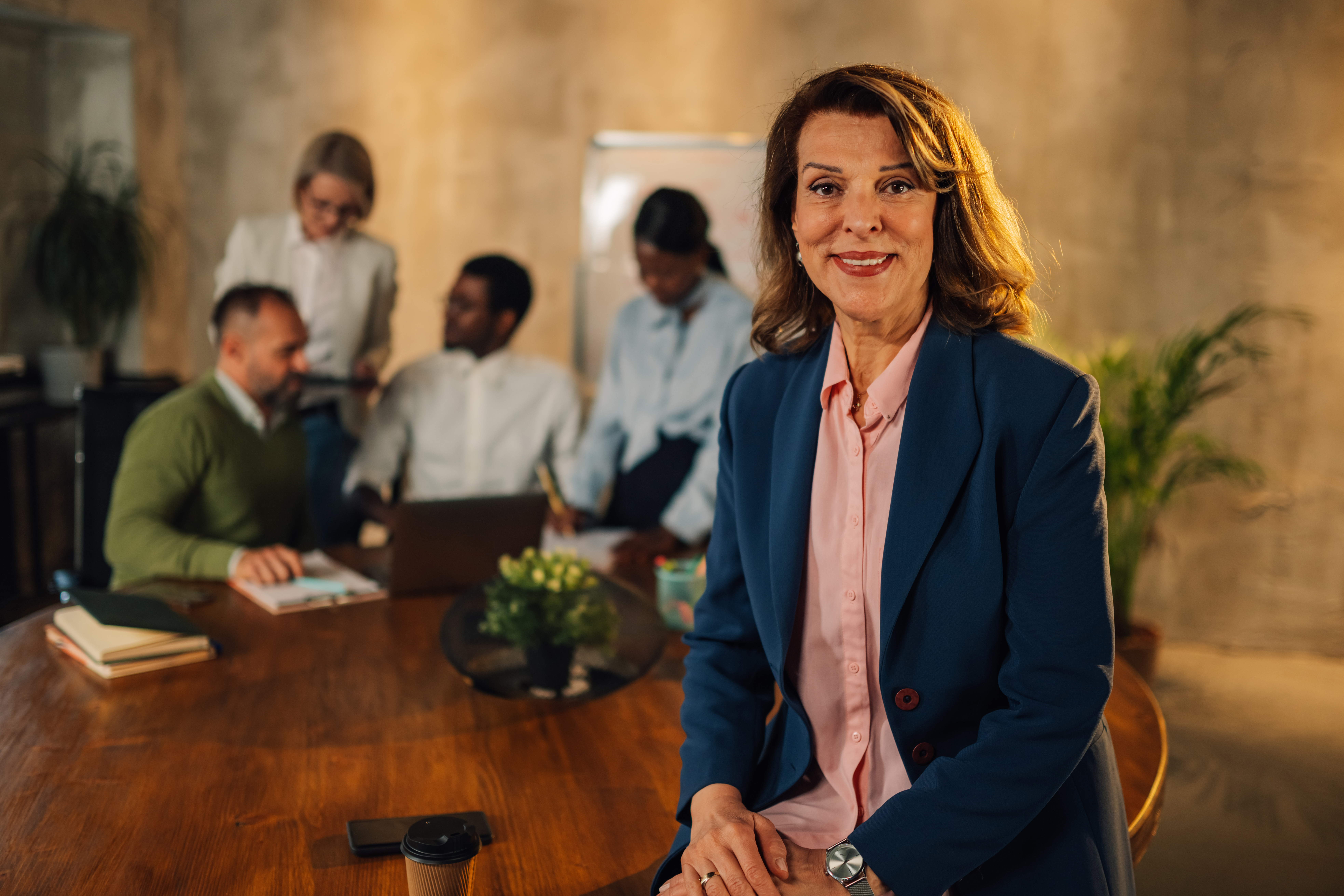 Portrait of a senior successful female manager sitting on a desk at modern conference room during the meeting of her multicultural staff. Confident mature businesswoman smiling at camera at office.